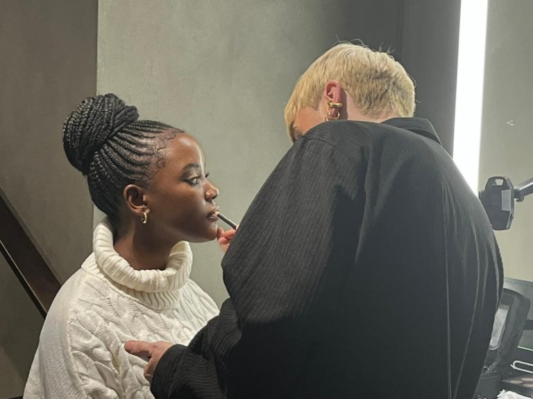 Black woman with braids, having her make up done by a man with short blond hair.