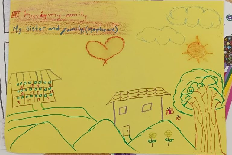 A picture of houses and trees, with a sun and clouds. The artist has written: "having my family" "My sister and family (nephews)"