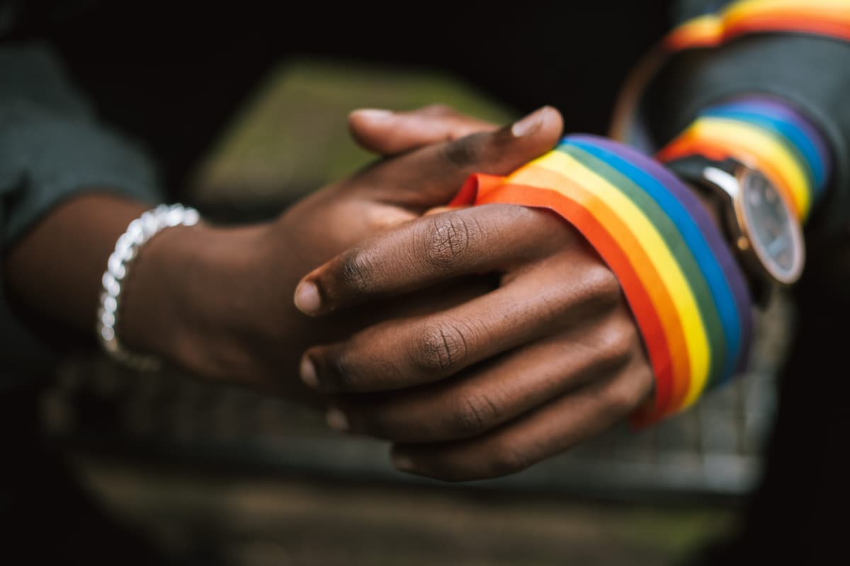 Hands of a black person clasped together, a rainbow ribbon wrapped around one