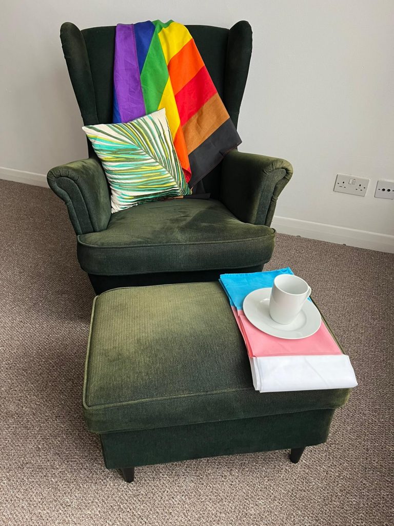 Green armchair and foot stool. There is a rainbow flag on the armchair, and a trans flag on the footstall